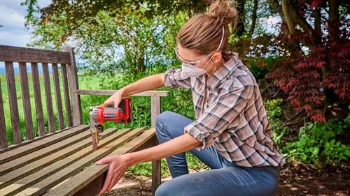 Woman using a jigsaw to cut a wooden bench outdoors.